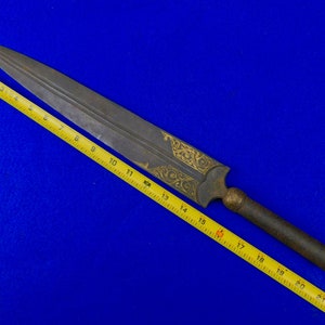 May include: A long, silver-colored dagger with a wooden handle and gold-colored decorative patterns. The dagger is measured against a yellow tape measure, showing it is approximately 19 inches long.