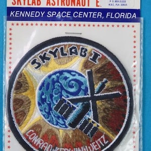 Puede incluir: Un parche bordado en blanco y negro con el texto "SKYLAB I" y "CONRAD-KERWIN WEITZ". El parche presenta una imagen estilizada de la estación espacial Skylab orbitando la Tierra. El parche está sobre un fondo blanco con estrellas rojas.