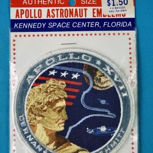 May include: A white and blue embroidered patch with the text "Apollo XVII" and the names "Cernan, Evans, Schmitt". The patch features a stylized image of a man's head with a blue and white background.