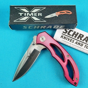 May include: A pink pocket knife with a silver blade and a black handle. The knife is open and the blade is visible. The knife is in a box with the brand name "Schrade" on it.