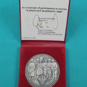May include: A silver-toned commemorative medal featuring five soldiers in historical uniforms, housed in a red velvet-lined case. The case lid displays text dedicated to Lt. General Ronald ADAMS for the exercise CONSTANT HARMONY 2000, signed by Ryszard ZUCHOWSKI.