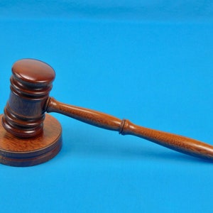 May include: A wooden gavel with a brown finish sits on a circular base. The gavel is lying on its side with the handle extending to the right.