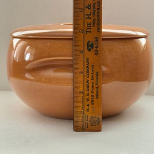 May include: A round, lidded ceramic bowl in a warm, earthy tone. The bowl is approximately 6 inches tall, with a matching lid. The surface has a glossy finish. The bowl is a decorative piece for the kitchen or dining room.