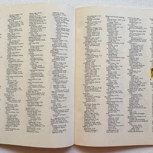 K&ouml;nnte beinhalten: Offene Buchseiten mit einer Liste von Rezepten und Zutaten. Der Text ist in schwarzer Schrift auf cremefarbenem Hintergrund. Eine kleine Illustration von zwei M&auml;dchen befindet sich in der Ecke. Das Buch ist auf einer wei&szlig;en Oberfl&auml;che ge&ouml;ffnet.