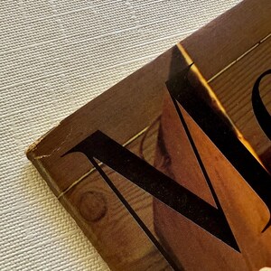 K&ouml;nnte beinhalten: Nahaufnahme eines Vintage-Buchcovers mit einem gro&szlig;en, stilisierten schwarzen Buchstaben "M" vor einem braunen Hintergrund mit Holzmaserung. Das Cover weist Gebrauchsspuren auf, die auf Alter hindeuten. Der Hintergrund ist eine strukturierte, cremefarbene Oberfl&auml;che.