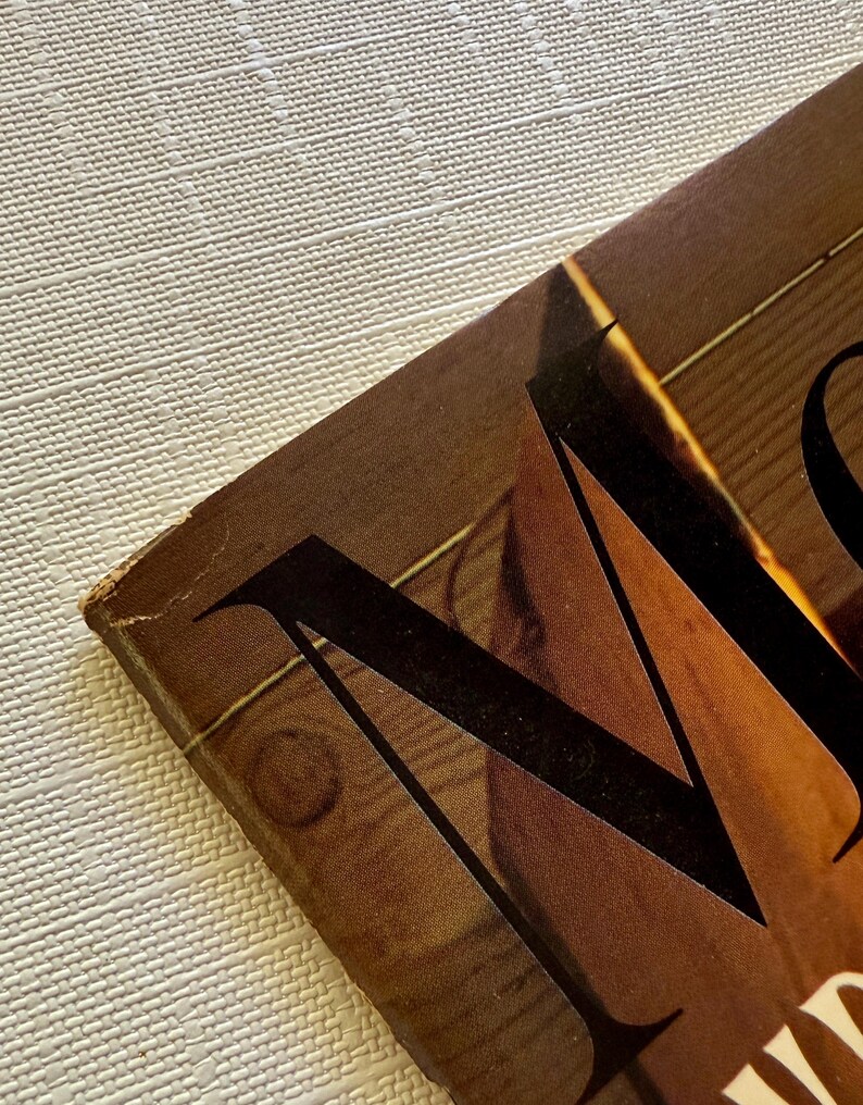 K&ouml;nnte beinhalten: Nahaufnahme eines Vintage-Buchcovers mit einem gro&szlig;en, stilisierten schwarzen Buchstaben "M" vor einem braunen Hintergrund mit Holzmaserung. Das Cover weist Gebrauchsspuren auf, die auf Alter hindeuten. Der Hintergrund ist eine strukturierte, cremefarbene Oberfl&auml;che.