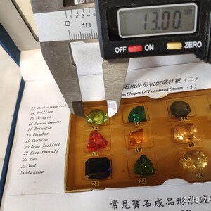 K&ouml;nnte beinhalten: Nahaufnahme einer Schieblehre, die einen gr&uuml;nen Edelstein misst. Die Schieblehre zeigt 13,00 an. Der Edelstein ist Teil einer Ausstellung verschiedener Edelsteinformen, die in Englisch und Chinesisch beschriftet sind.