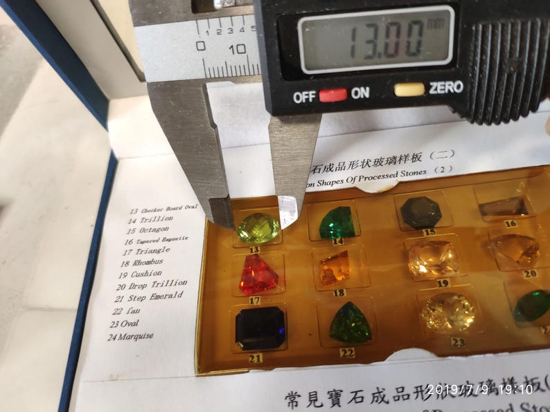 K&ouml;nnte beinhalten: Nahaufnahme einer Schieblehre, die einen gr&uuml;nen Edelstein misst. Die Schieblehre zeigt 13,00 an. Der Edelstein ist Teil einer Ausstellung verschiedener Edelsteinformen, die in Englisch und Chinesisch beschriftet sind.