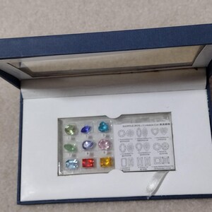 K&ouml;nnte beinhalten: Eine blaue Samtbox mit wei&szlig;em Innenraum. Die Box enth&auml;lt ein kleines Plastiktablett mit 12 bunten Edelsteinen und eine Karte mit einem Diagramm verschiedener Edelsteinschliffe. Die Karte lautet "SAMPLE STONES - Gemstone Cuts".