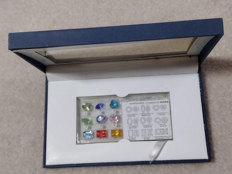 K&ouml;nnte beinhalten: Eine blaue Samtbox mit wei&szlig;em Innenraum. Die Box enth&auml;lt ein kleines Plastiktablett mit 12 bunten Edelsteinen und eine Karte mit einem Diagramm verschiedener Edelsteinschliffe. Die Karte lautet "SAMPLE STONES - Gemstone Cuts".