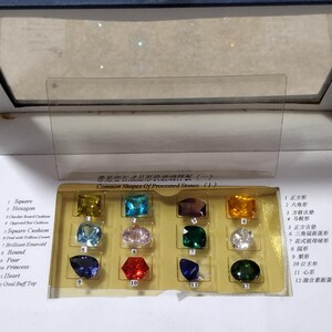 K&ouml;nnte beinhalten: Eine Vitrine mit 12 verschiedenen Edelsteinsformen, darunter quadratisch, sechseckig, quadratisches Kissen, brillanter Smaragd, rund, Birne, Prinzessin, Herz und ovaler Buff Top. Die Vitrine ist mit "H&auml;ufige Formen von bearbeiteten Steinen" beschriftet.