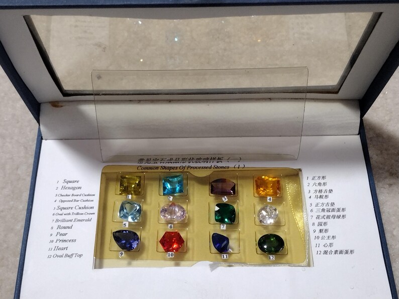 K&ouml;nnte beinhalten: Eine Vitrine mit 12 verschiedenen Edelsteinsformen, darunter quadratisch, sechseckig, quadratisches Kissen, brillanter Smaragd, rund, Birne, Prinzessin, Herz und ovaler Buff Top. Die Vitrine ist mit "H&auml;ufige Formen von bearbeiteten Steinen" beschriftet.