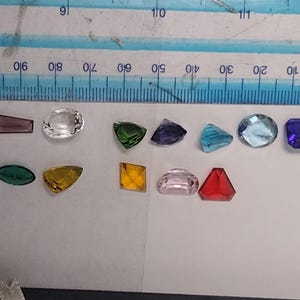 K&ouml;nnte beinhalten: Sortiment aus bunten, facettierten Glas-Edelsteinen in verschiedenen Formen und Gr&ouml;&szlig;en, darunter ein klarer Oval, ein gr&uuml;nes Dreieck, ein violettes Quadrat, ein blauer Oval, ein rotes Dreieck, ein rosa Oval, ein gelbes Dreieck, ein gr&uuml;ner Oval, ein violettes Rechteck und ein blaues Rechteck.