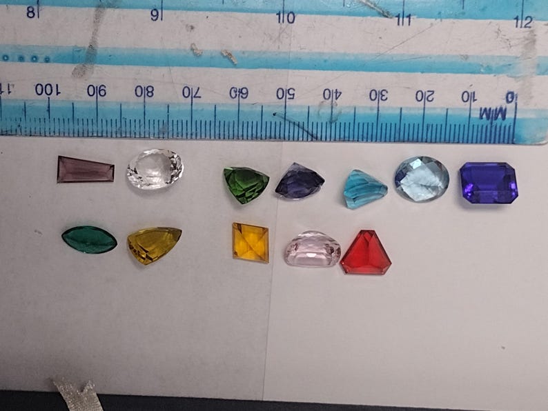 K&ouml;nnte beinhalten: Sortiment aus bunten, facettierten Glas-Edelsteinen in verschiedenen Formen und Gr&ouml;&szlig;en, darunter ein klarer Oval, ein gr&uuml;nes Dreieck, ein violettes Quadrat, ein blauer Oval, ein rotes Dreieck, ein rosa Oval, ein gelbes Dreieck, ein gr&uuml;ner Oval, ein violettes Rechteck und ein blaues Rechteck.