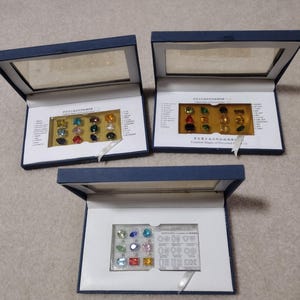 K&ouml;nnte beinhalten: Drei blaue Samtboxen mit durchsichtigen Deckeln, die kleine, bunte Edelsteine enthalten. Die Boxen sind mit dem Text "H&auml;ufige Formen von Edelsteinen" beschriftet.