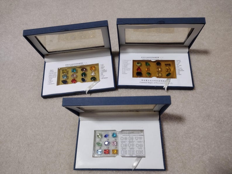 K&ouml;nnte beinhalten: Drei blaue Samtboxen mit durchsichtigen Deckeln, die kleine, bunte Edelsteine enthalten. Die Boxen sind mit dem Text "H&auml;ufige Formen von Edelsteinen" beschriftet.