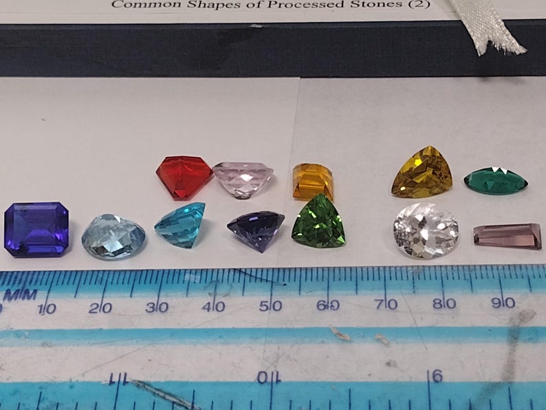 K&ouml;nnte beinhalten: Ein Lineal mit Markierungen in Millimetern und Zoll, auf dem zehn verschiedene Edelsteinform in einer Reihe liegen. Die Edelsteine sind in verschiedenen Farben, darunter rot, rosa, gelb, gr&uuml;n, blau und lila.