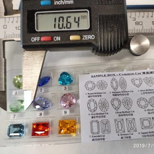 K&ouml;nnte beinhalten: Eine Nahaufnahme einer Schieblehre, die einen kleinen, klaren Edelstein misst. Die Schieblehre zeigt 10,64 mm an. Der Edelstein ist einer von vielen in einer Musterbox mit der Aufschrift "SAMPLE BOX - Common Cut 常見宝石". Die Box enth&auml;lt verschiedene Formen von Edelsteinen, darunter runde, ovale, birnenf&ouml;rmige, herzf&ouml;rmige und quadratische.