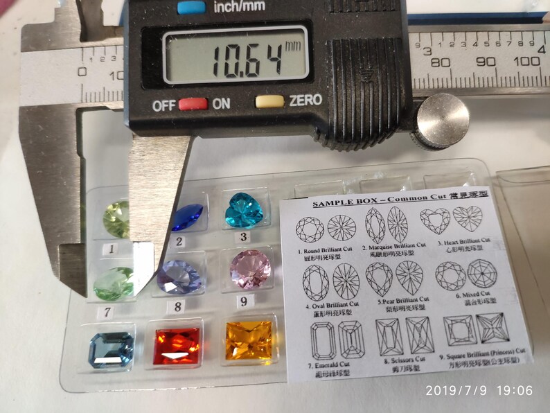 K&ouml;nnte beinhalten: Eine Nahaufnahme einer Schieblehre, die einen kleinen, klaren Edelstein misst. Die Schieblehre zeigt 10,64 mm an. Der Edelstein ist einer von vielen in einer Musterbox mit der Aufschrift "SAMPLE BOX - Common Cut 常見宝石". Die Box enth&auml;lt verschiedene Formen von Edelsteinen, darunter runde, ovale, birnenf&ouml;rmige, herzf&ouml;rmige und quadratische.