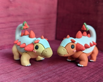 May include: Two small, hand-painted ceramic dinosaur figurines with a strawberry theme. The dinosaurs are orange and yellow with white and blue accents. They have a strawberry design on their backs and a strawberry on their tails.