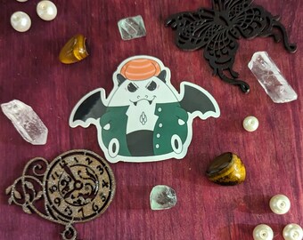 May include: A sticker of a cartoon vampire in a green jacket and orange hat, with a black butterfly, pearls, crystals, and a clock. These decorative items are arranged on a dark red wooden surface.