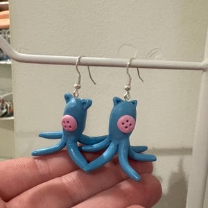May include: A pair of handmade blue octopus earrings with pink accents. Each earring features a small octopus figure with eight tentacles and a pink face. The earrings are suspended from silver-colored fish hook ear wires.