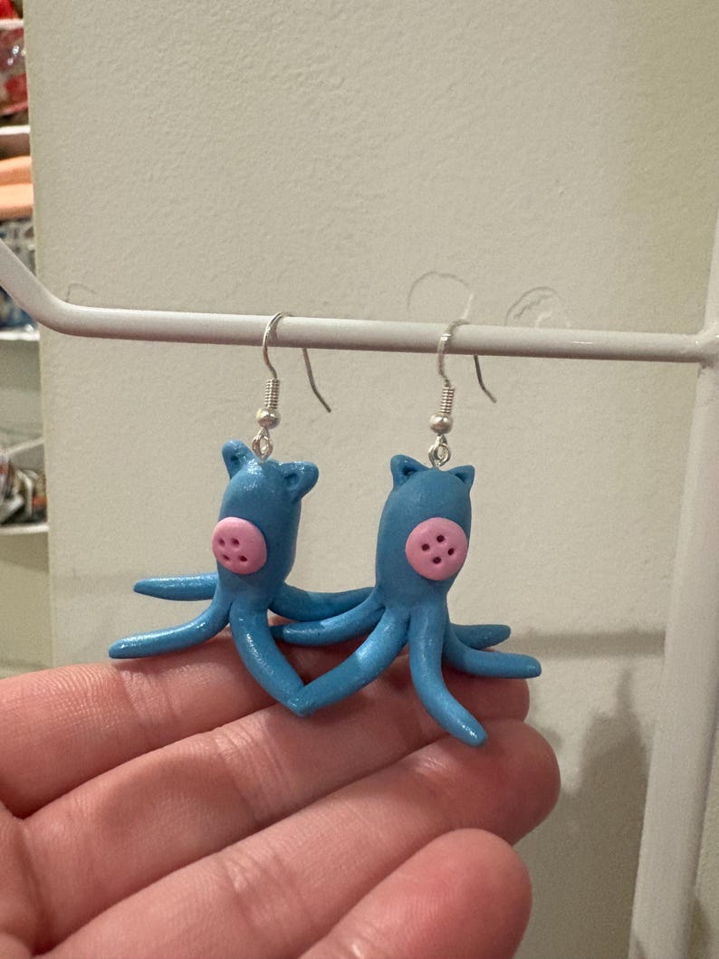 May include: A pair of handmade blue octopus earrings with pink accents. Each earring features a small octopus figure with eight tentacles and a pink face. The earrings are suspended from silver-colored fish hook ear wires.