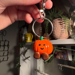 May include: A cheerful orange pumpkin keychain with a green stem and brown feet. The pumpkin has a smiling face and is attached to a silver metal keyring and clasp. A fun, seasonal accessory.