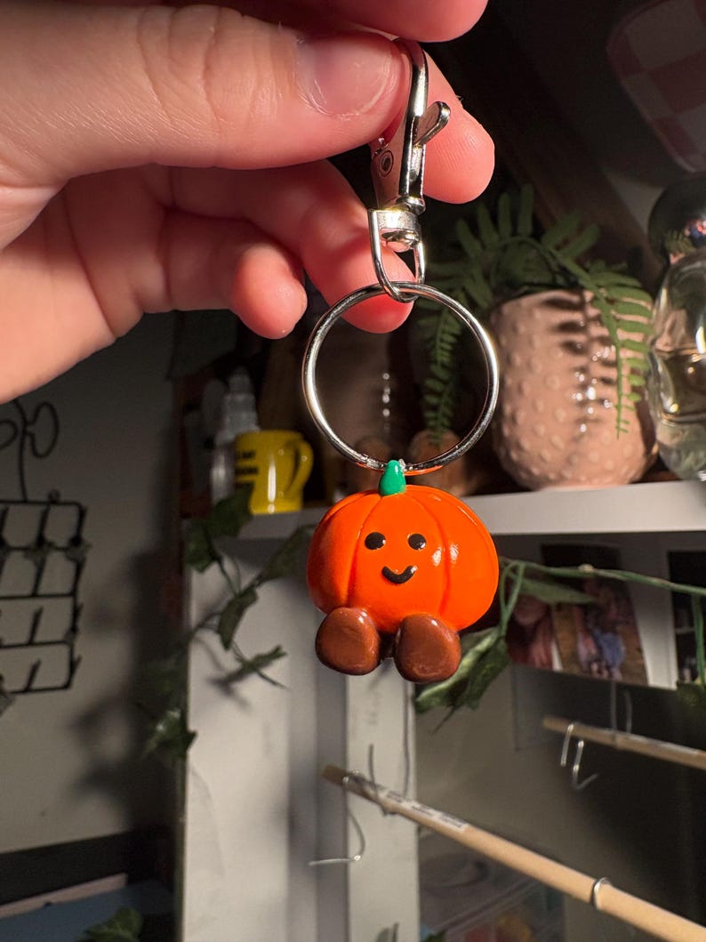 May include: A cheerful orange pumpkin keychain with a green stem and brown feet. The pumpkin has a smiling face and is attached to a silver metal keyring and clasp. A fun, seasonal accessory.