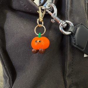 May include: A whimsical orange pumpkin charm with a smiling face, green stem, and brown feet hangs from a gold-colored keychain. The charm is attached to a black tote bag with a silver clasp.