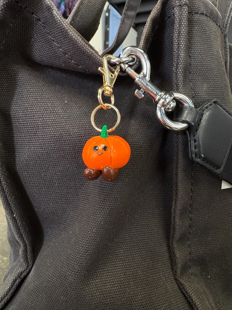 May include: A whimsical orange pumpkin charm with a smiling face, green stem, and brown feet hangs from a gold-colored keychain. The charm is attached to a black tote bag with a silver clasp.