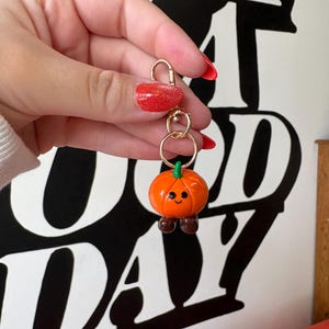 May include: A gold-colored keychain with a small, orange pumpkin charm. The pumpkin has a green stem, brown feet, and a smiling face. The keychain is held up by a hand with red nail polish.