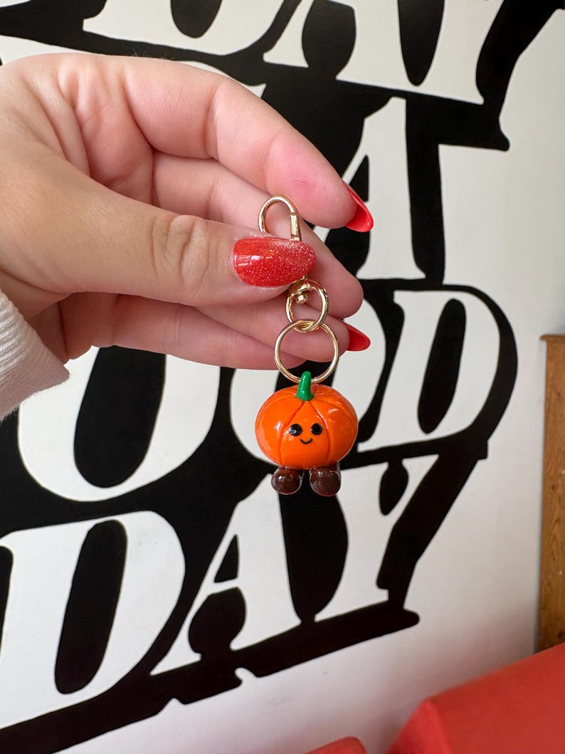 May include: A gold-colored keychain with a small, orange pumpkin charm. The pumpkin has a green stem, brown feet, and a smiling face. The keychain is held up by a hand with red nail polish.