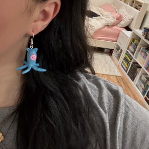 May include: A light blue octopus-shaped earring with pink accents and silver-colored ear wire. The earring is a whimsical, handmade accessory. The octopus has eight tentacles and a round head with two pink buttons.