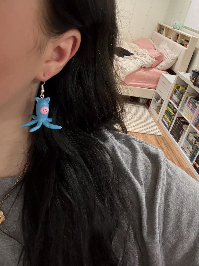 May include: A light blue octopus-shaped earring with pink accents and silver-colored ear wire. The earring is a whimsical, handmade accessory. The octopus has eight tentacles and a round head with two pink buttons.