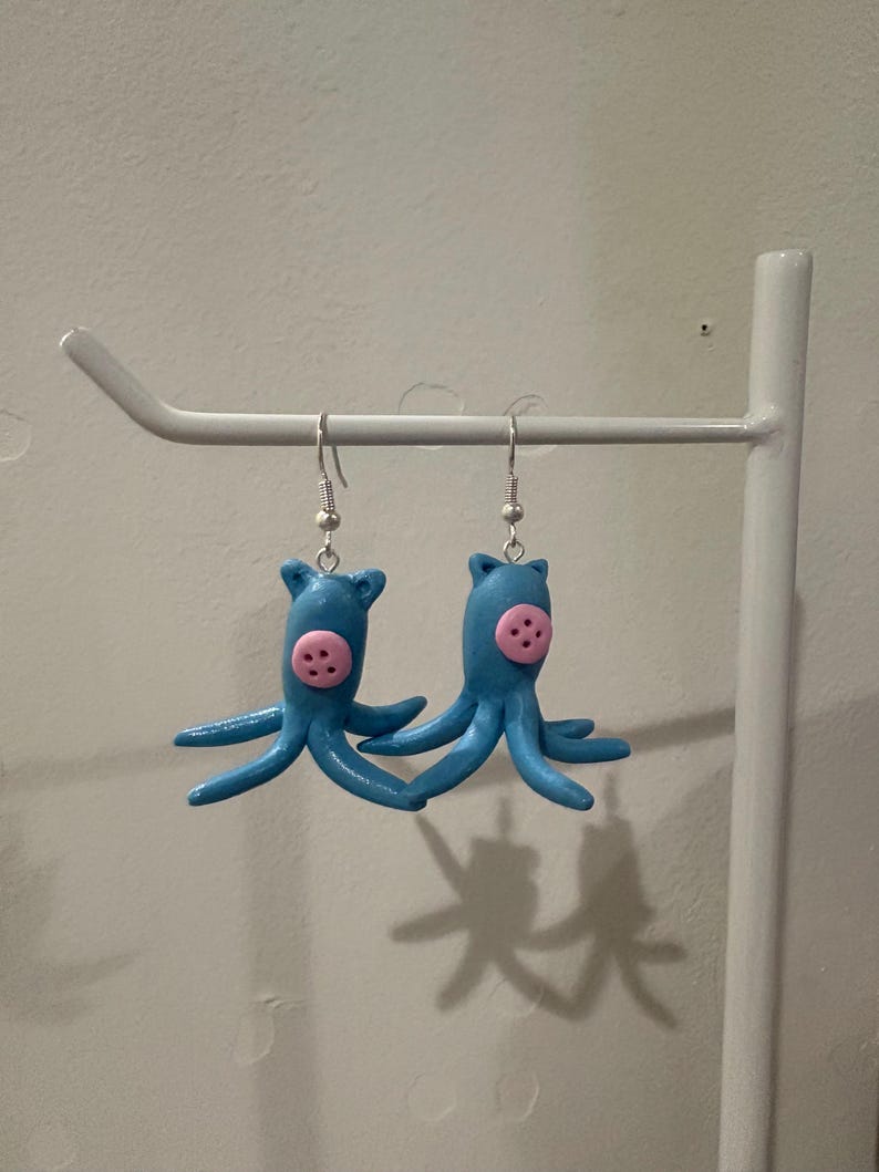 May include: A pair of handmade blue octopus earrings with pink accents. Each earring features a small, round pink button and multiple blue tentacles. The earrings are suspended from silver-colored fish hook ear wires.