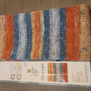 May include: Close-up of a knitted sock with a color gradient of orange, blue, and cream. The sock is labeled "Yarn Bee COZY TOES" with the color name "Soft Dusk". The sock is made of a soft, textured yarn.