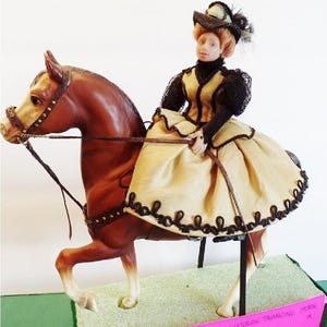 May include: A brown horse with a black bridle and saddle. A doll wearing a tan dress with black lace trim and a black hat is riding the horse. The horse is on a stand with a pink sign that reads '1963 'CHEYENNE' WESTERN PRANCING HORSE, SIDE SADDLE, BRIDLE,  $35.00.'