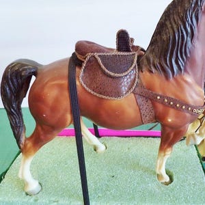 May include: A brown horse figurine with a detailed saddle. The horse has a dark brown mane and tail, and the saddle is a textured brown with decorative stitching. The figurine is set on a green surface.