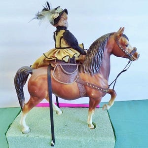 May include: A brown horse figurine with a rider in a yellow and black outfit and a feathered hat. The horse has a saddle and bridle. The figurine is on a green base.