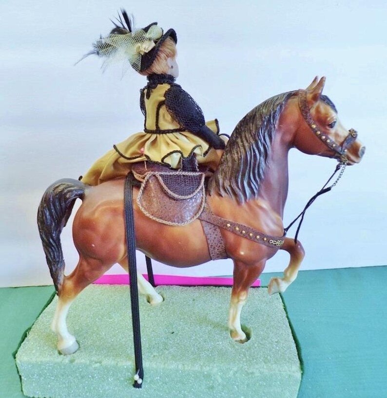 May include: A brown horse figurine with a rider in a yellow and black outfit and a feathered hat. The horse has a saddle and bridle. The figurine is on a green base.
