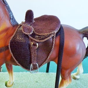 May include: A brown horse figurine with a detailed saddle. The saddle is dark brown with a textured pattern and gold accents. The horse has a brown coat and a black mane and tail.