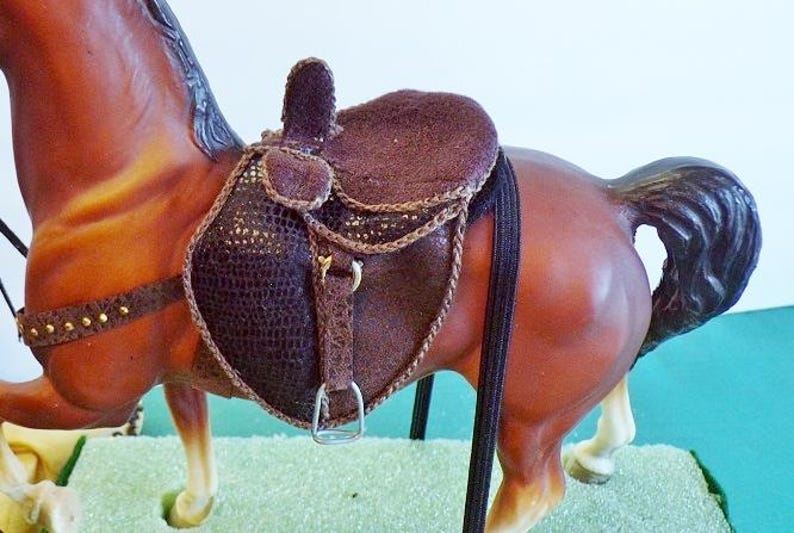 May include: A brown horse figurine with a detailed saddle. The saddle is dark brown with a textured pattern and gold accents. The horse has a brown coat and a black mane and tail.