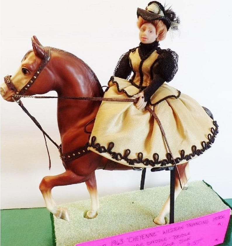 May include: A brown horse with a black bridle and saddle. A doll wearing a tan dress with black lace trim and a black hat is riding the horse. The horse is on a stand with a pink sign that reads '1963 'CHEYENNE' WESTERN PRANCING HORSE, SIDE SADDLE, BRIDLE,  $35.00.'