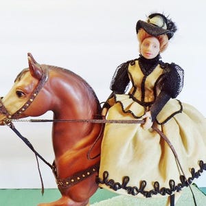 May include: A doll depicting a woman riding a brown horse. The woman is dressed in a yellow gown with black trim and a black hat. The horse has a detailed bridle and reins.