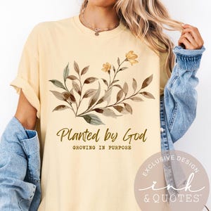 May include: Cream-colored t-shirt with the text "Planted by God" and a floral design in brown and yellow. The shirt also has the text "Growing in Purpose" below the main text.