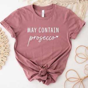 May include: A mauve t-shirt with the text "MAY CONTAIN prosecco" in white lettering with a small heart symbol.
