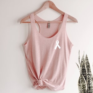 May include: A pink tank top with a white ribbon on the front. The tank top is hanging on a wooden hanger.