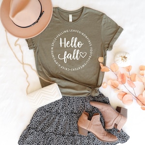 May include: An olive green t-shirt with the words "Hello Fall" in a circular design, surrounded by fall-themed words. The shirt is paired with a black and white patterned skirt, tan ankle boots, a tan hat, and a white purse with a gold chain.