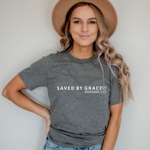 May include: A gray t-shirt with the text "SAVED BY GRACE" and a heart symbol. Below the text is "EPHESIANS 2:10".